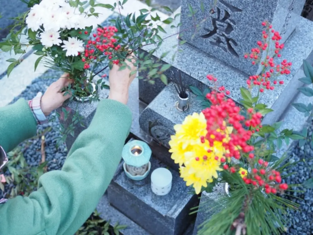 墓前に季節の花を供える人の手元と、線香やロウソクが並ぶお参りの場面