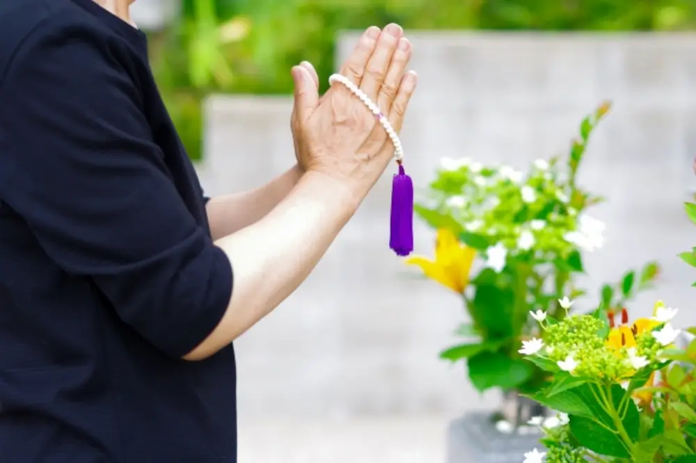 数珠を持った高齢の方が花の前で手を合わせて祈る姿のクローズアップ写真