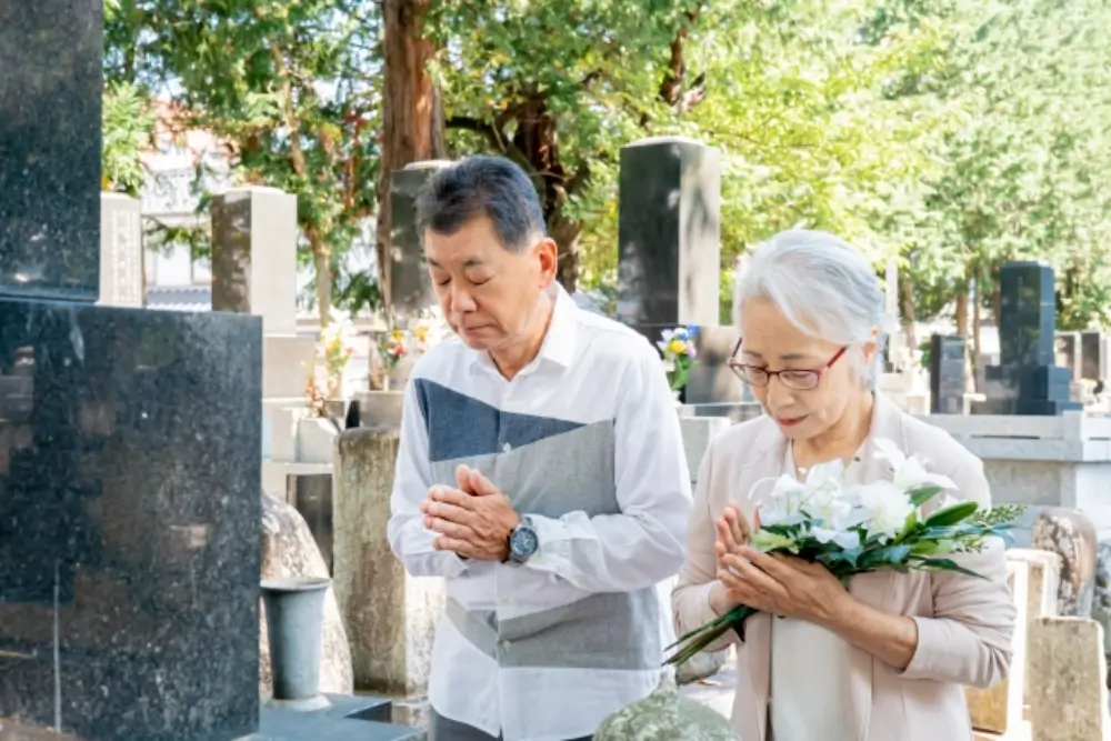 高齢の夫婦が白い花を手に持ち、墓前で手を合わせて祈っている様子