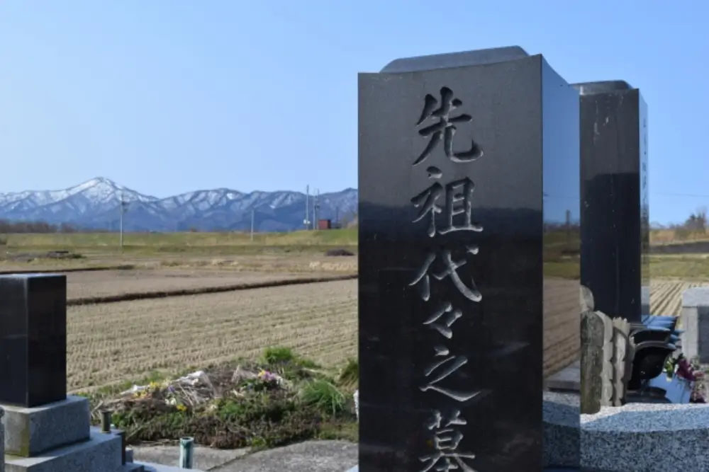 「先祖代々之墓」と刻まれた黒い墓石と山並みが見える田園風景