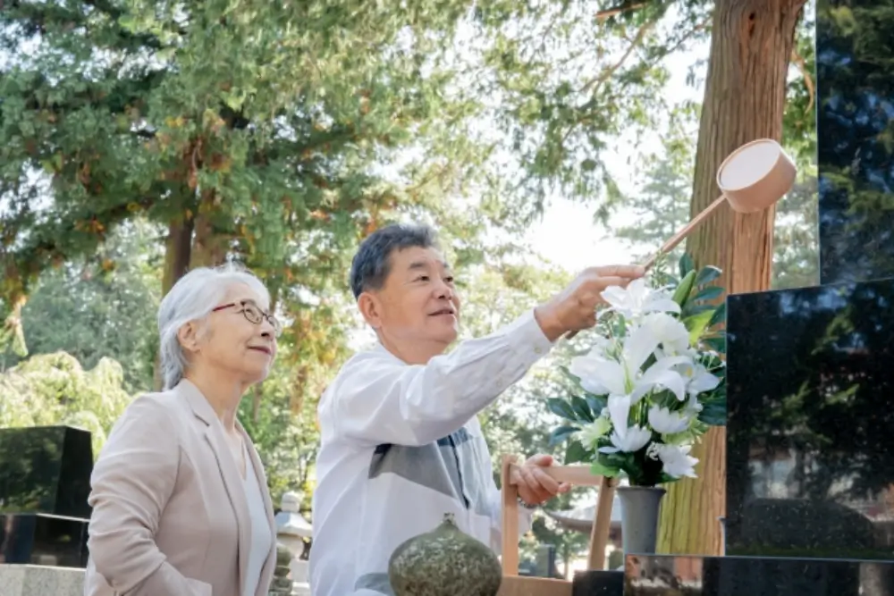 柄杓で墓石に水をかける高齢の男性と見守る女性、供養の風景
