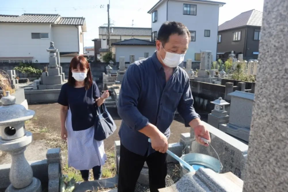 墓石を掃除する男性と見守る女性、マスク姿の夫婦の墓参り風景