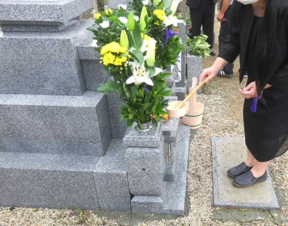 墓石に柄杓で水をかけて供養する喪服姿の女性と供えられた花々