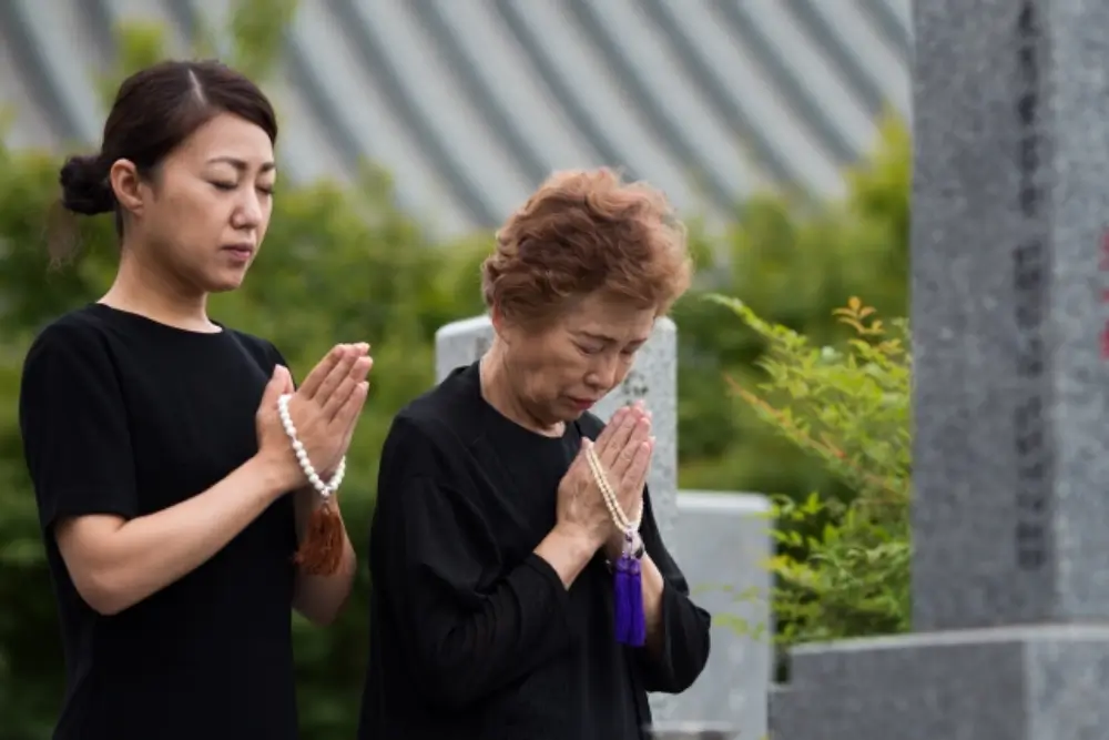 喪服の女性二人が墓前で数珠を持ち、心を込めて手を合わせて祈っている様子