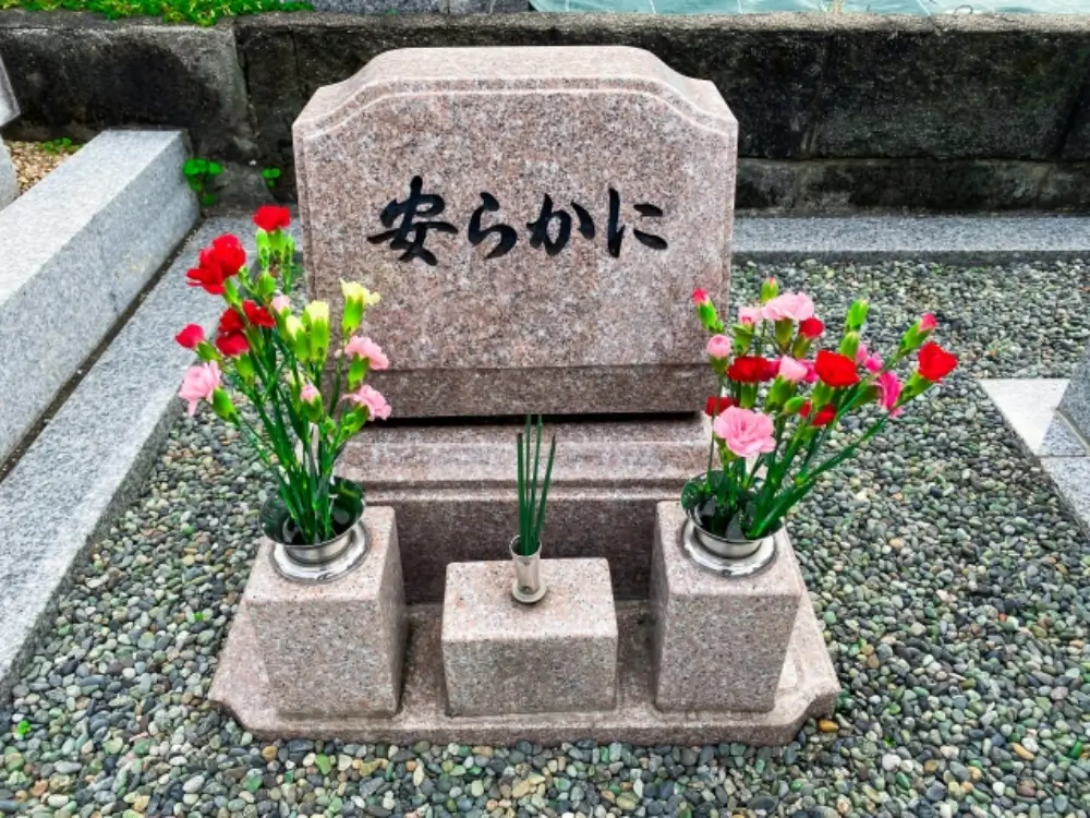 「安らかに」と刻まれたピンク色の墓石と供えられたカーネーションの花々